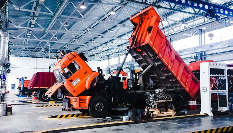 Ein orangefarbener Kipper mit gekipptem Fahrerhaus und angehobener Ladefläche in einer großen industriellen Werkstatt.